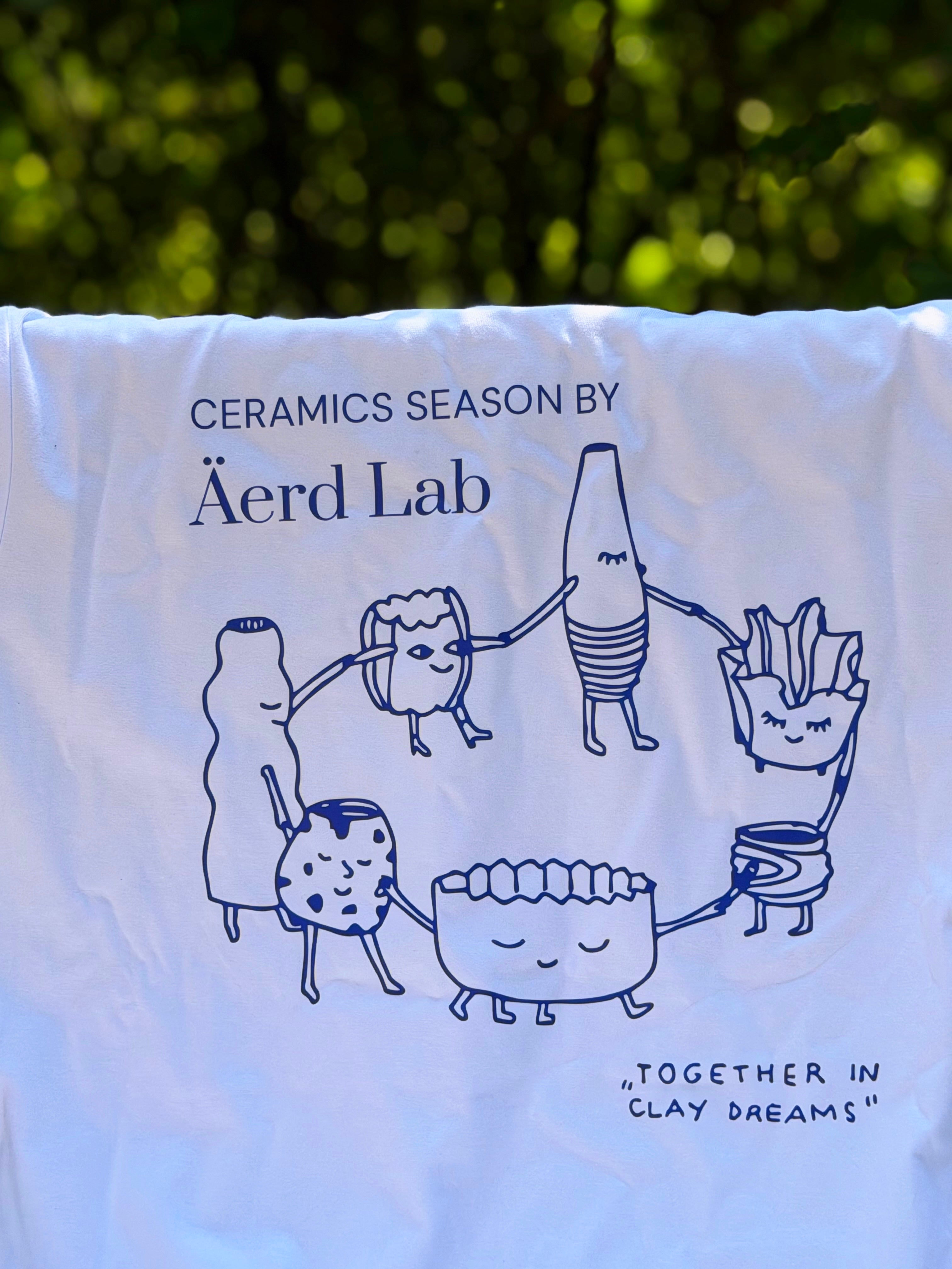 A white fabric item, likely a towel or blanket, printed with blue line art and text reading 'CERAMICS SEASON BY Äerd Lab' and 'TOGETHER IN CLAY DREAMS'. The fabric is hanging outdoors with a blurred green foliage background.